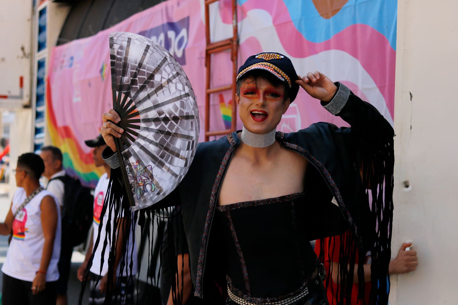 Drag queen com leque em frente ao backdrop da Parada